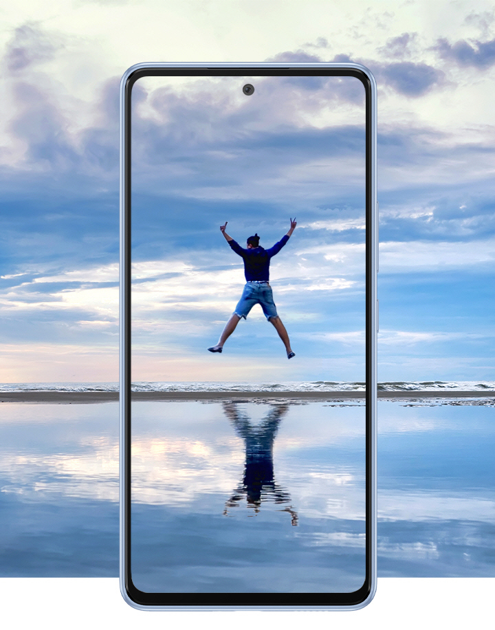 Galaxy A53 5G seen from the front against a beautiful landscape that overlaps onto the screen. It shows an expansive sky over water that reflects it with a thin horizon dissecting near the middle. In the center of the screen, a man is jumping in the air with all four limps outstretched and his reflection is shown on the water.