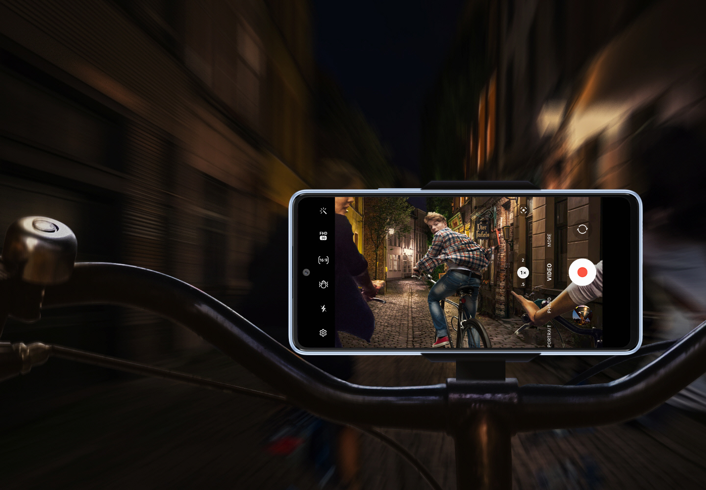 A frontal view of a Galaxy A53 5G mounted horizontally onto a bike handlebar. While the background is blurry and dark, the smartphone screen shows that it is clearly and stably recording what is happening: A group of bike riders are riding through a dimly lit road at night, with the rider furthest ahead looking back at the phone.