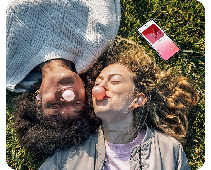 Two female friends, both wearing Buds Pro, are lying down on the grass on a sunny day, blowing bubbles with bubble gum. A smartphone laid down nearby shows a song being played, illustrating that both friends are listening to the song together with their Buds products.