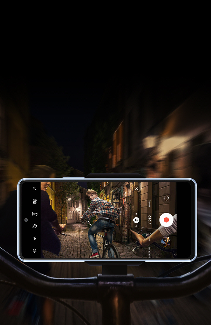 A frontal view of a Galaxy A53 5G mounted horizontally onto a bike handlebar. While the background is blurry and dark, the smartphone screen shows that it is clearly and stably recording what is happening: A group of bike riders are riding through a dimly lit road at night, with the rider furthest ahead looking back at the phone.