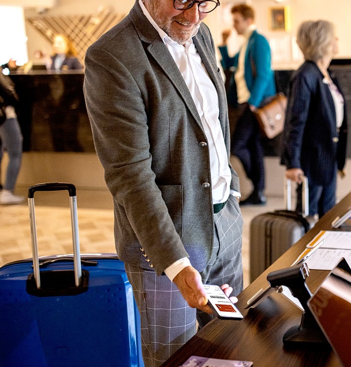An old man with a luggage is standing in the middle of the picture, holding the phone with Samsung Wallet card on screen. He is standing in front of the card reader. 